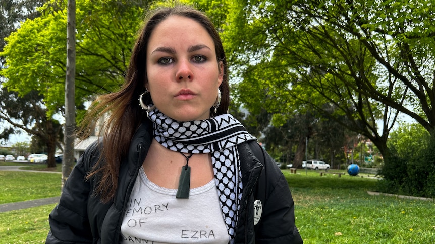 Aaneke sits in a park with brown hair, hoop earrings, black and white scarf, black puffer and white singlet with names on it. 