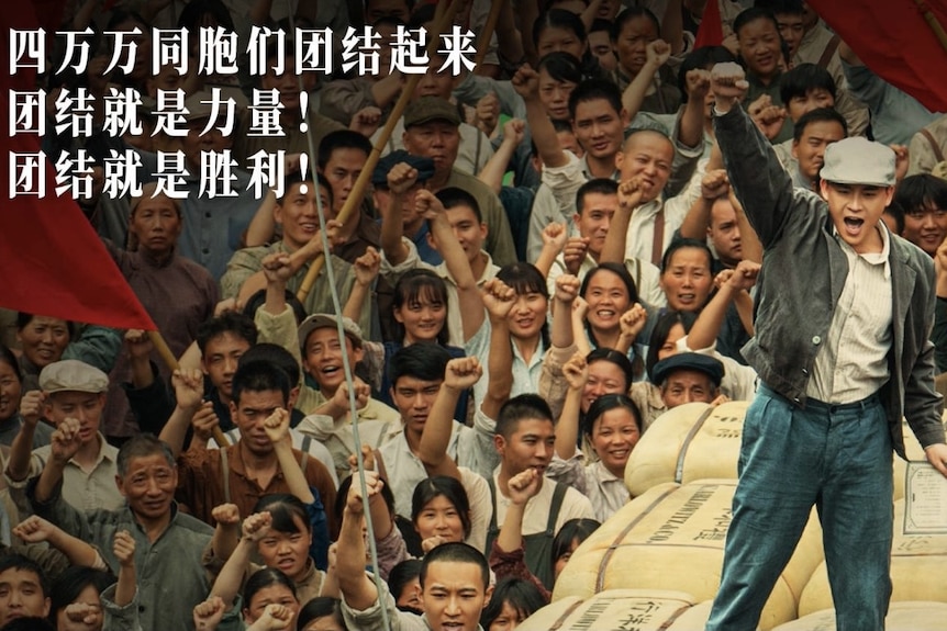 A young men stands on a pile of gunny bags, raising his right arm. Crowds follow him.  