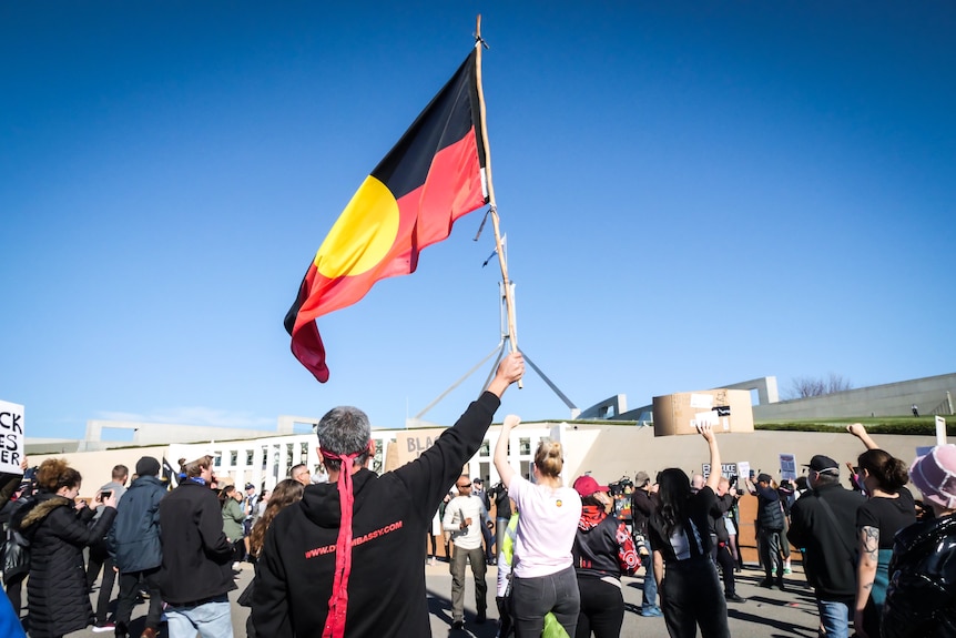 A group of protestors marching towards parliament house in Canberra, one waving an aboriginal flag high.