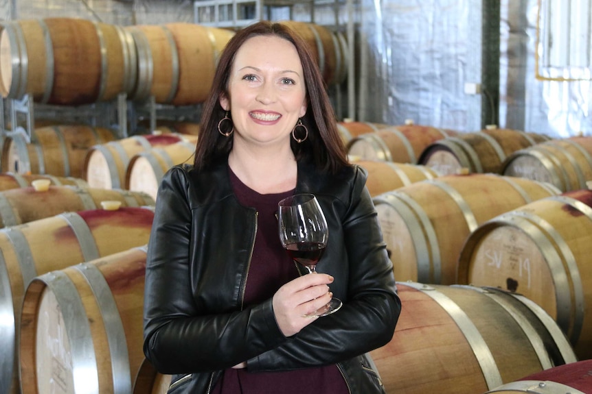 A smiling woman stands with a glass of wine in front of a row of wine barrels in a warehouse.