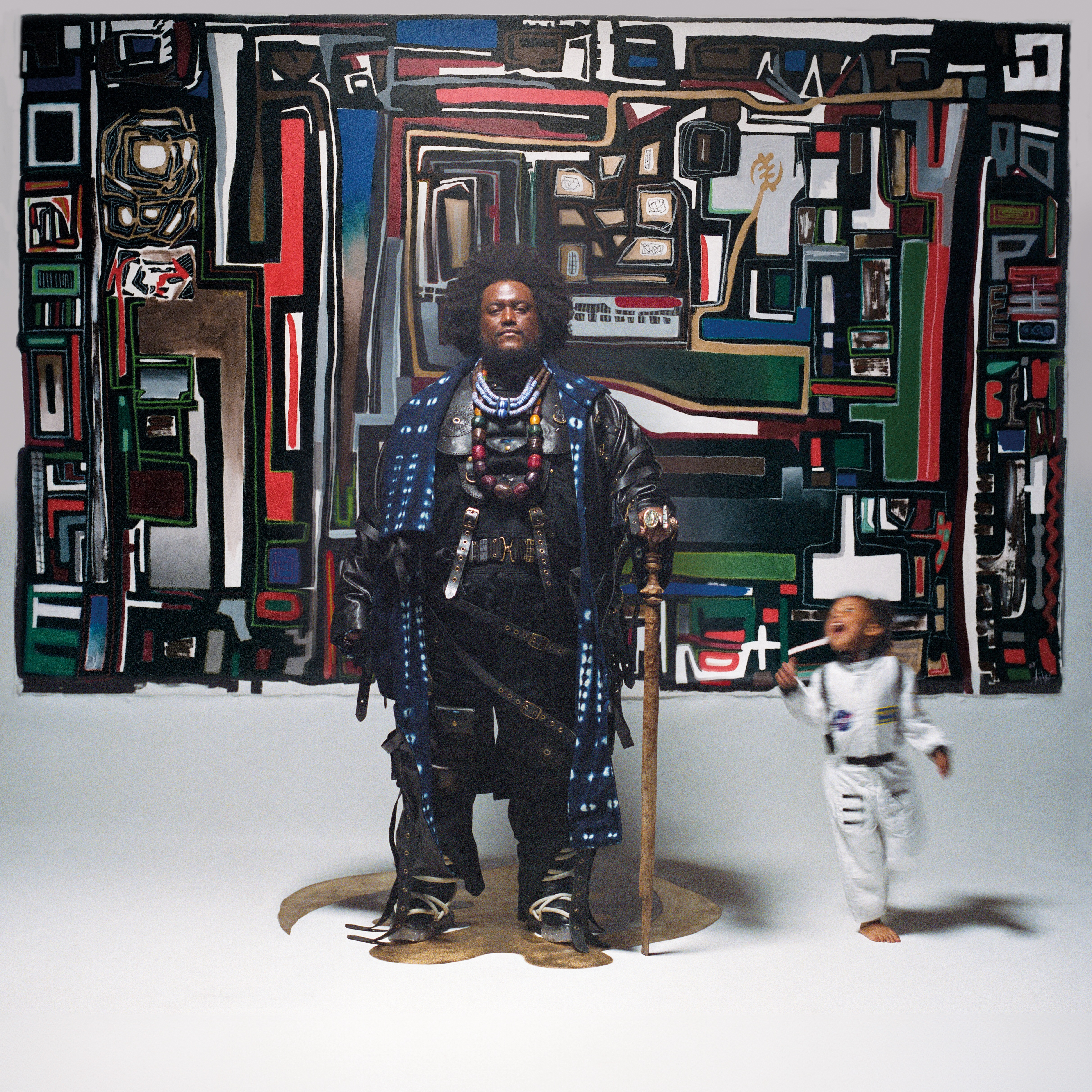 Kamasi Washington stands in front of a painted mural, wearing fusion Afro/samurai armour with his daughter running around