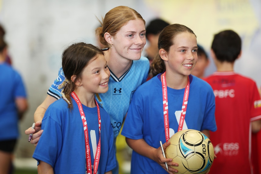 Sydney FC soccer player Cortnee Vine smiles for a photo with two kids. Cortnee is in a light blue Sydney FC shirt.