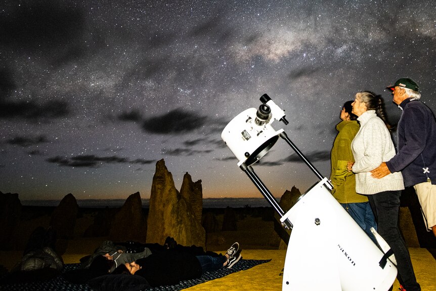 People look up at the sky next to a big telescope, rocky outcrops in the background.