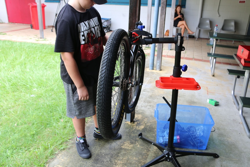 A boy standing in front of a partly disassembled bicycle hanging off the ground