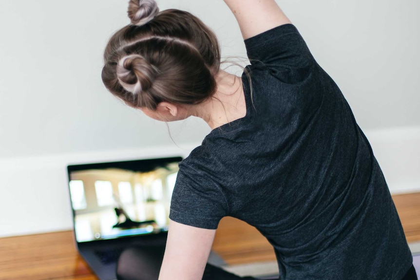 A woman sitting on a yoga mat stretches while facing an instructional video on a laptop.