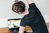 A woman sitting on a yoga mat stretches while facing an instructional video on a laptop.
