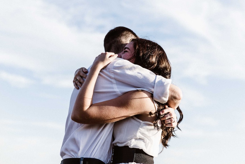 A man and woman hug outside, a blue and white sky beyond them.