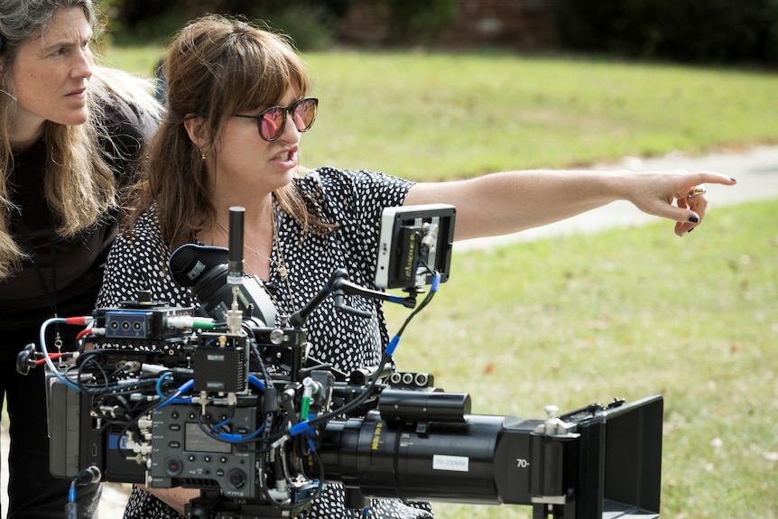 A woman in her 50s in a spotty dress and sunglasses sits behind a film camera and points, another blonde woman behind her