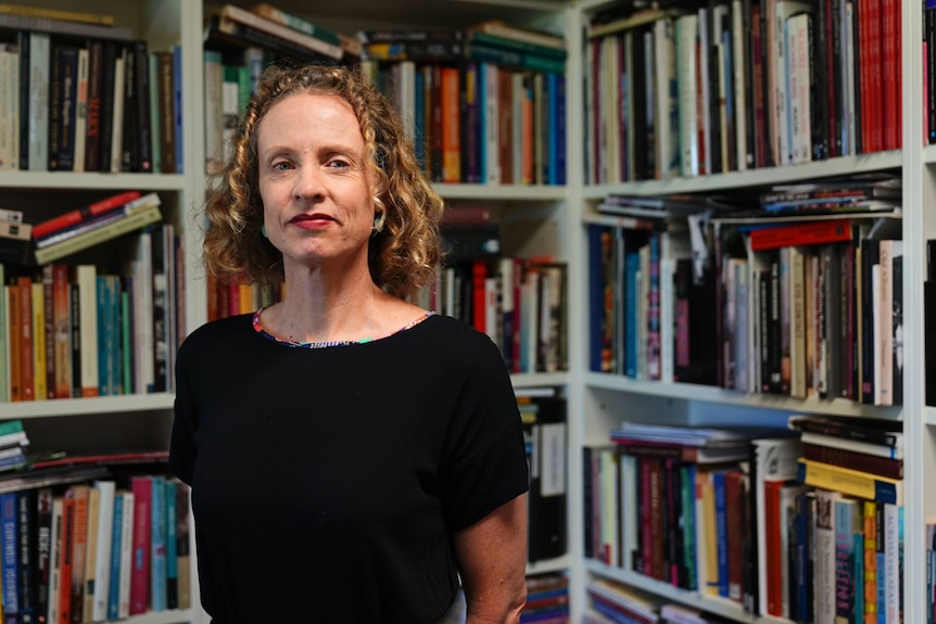 A woman with blonde curly hair stands in front of a bookshelf