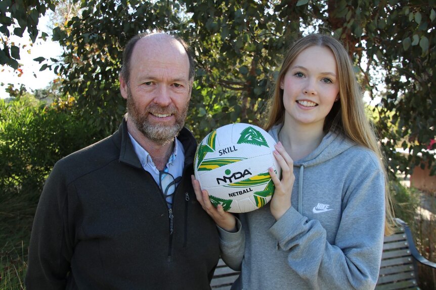 A teen age girl holds a netball and stands next to her father.