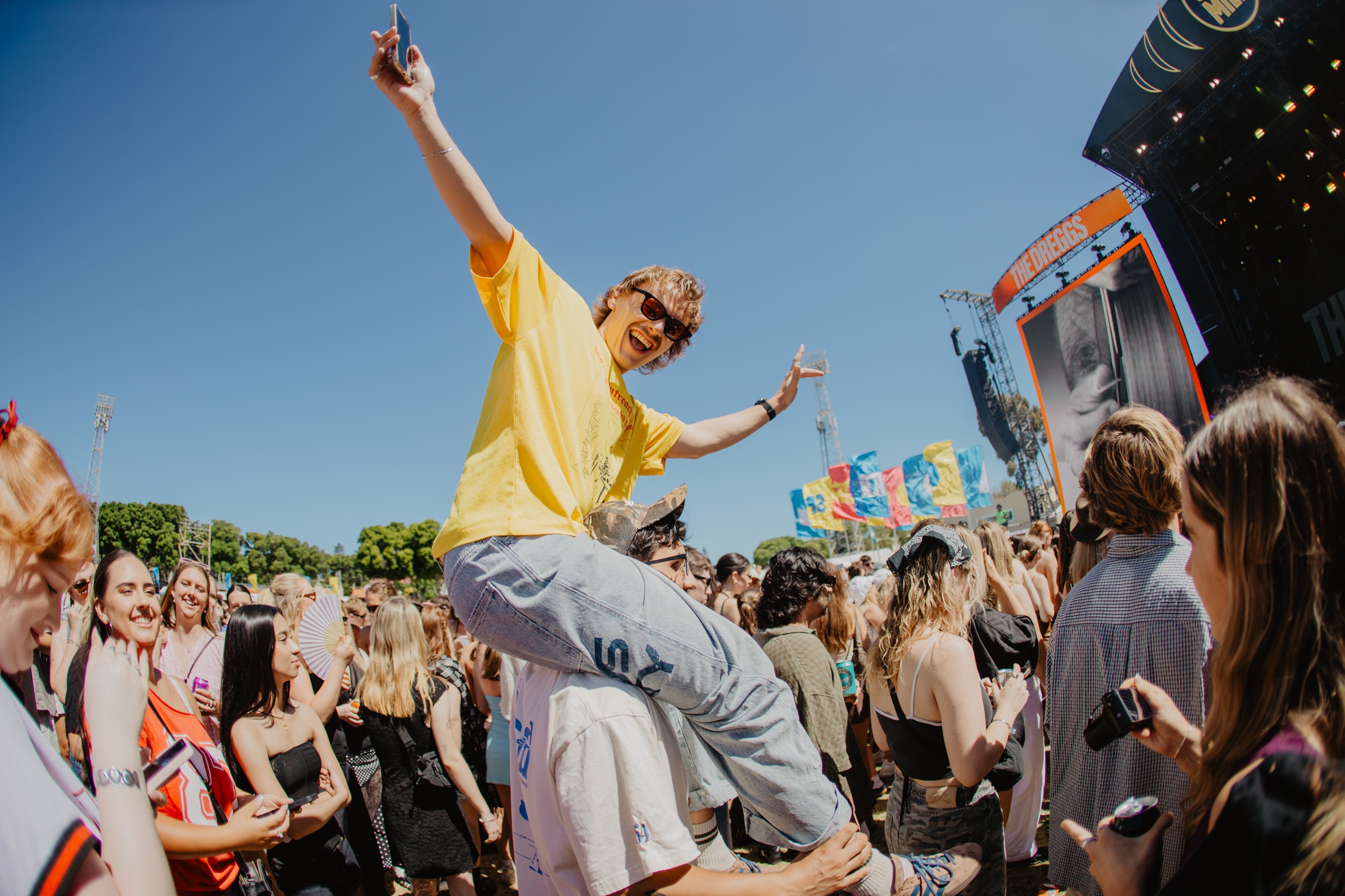 A man on the shoulders of someone else at a music festival with a crowd of people around him