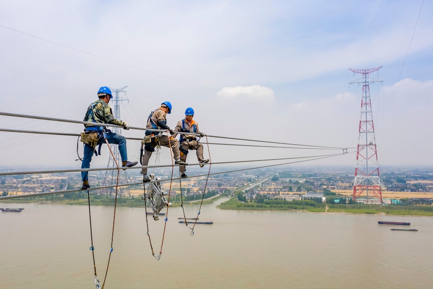 Three workers on long wires strung over water to a pylon in the distance.