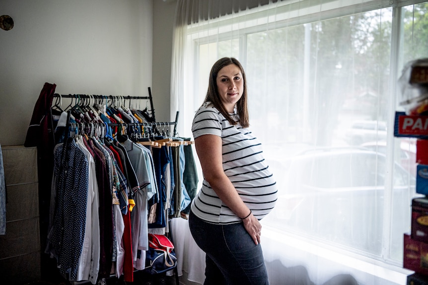 Jen stands near a window inside her living room, heavily pregnant. There are many clothes on hangers behind her. 
