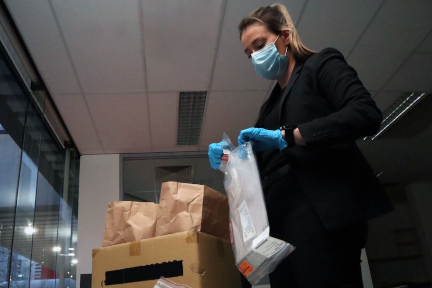 A woman in a black suit, face mask and gloves sorts through paper bags of evidence