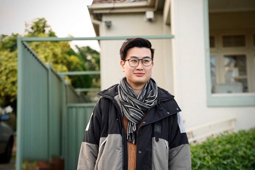 David Le stands with glasses and a scarf outside a house.