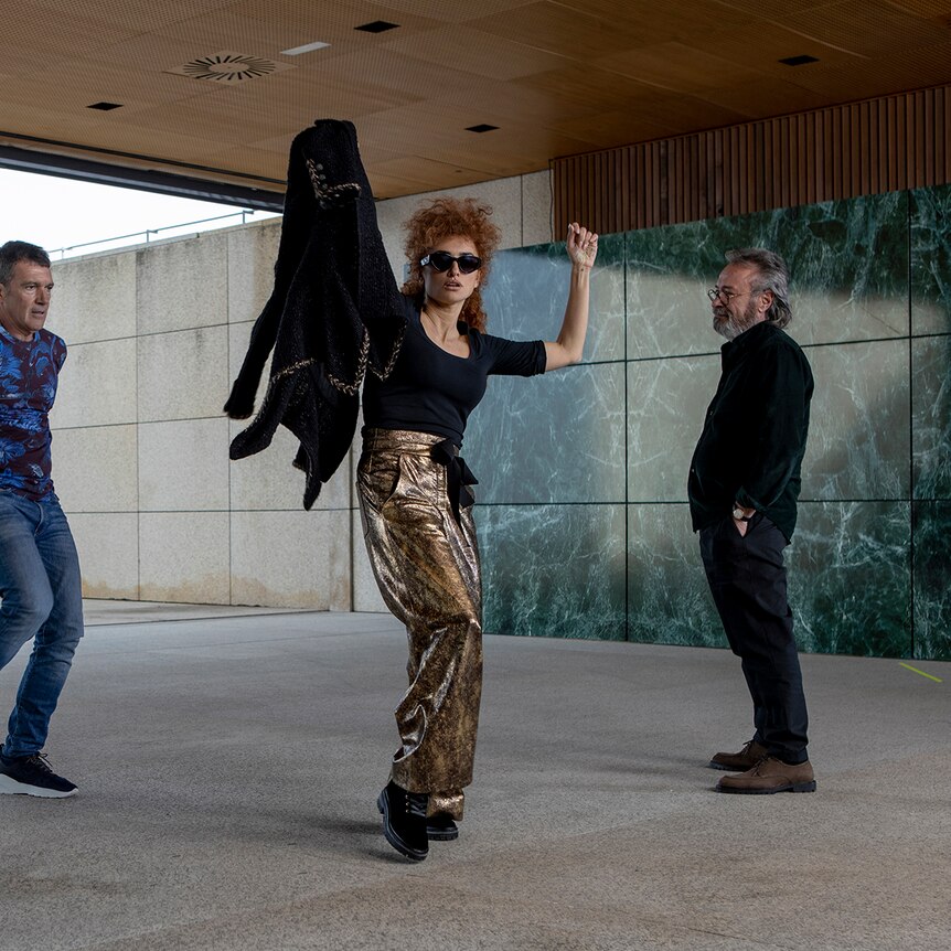 woman in sunglasses and gold trousers dancing while a man in black clothes watches and a man removes his blue jacket