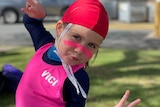 A young girl in a blue stinger suit, red cap and bright pink swim vest with pink zinc on her face.
