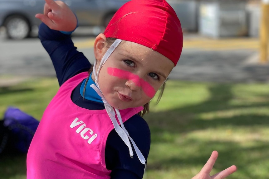 A young girl in a blue stinger suit, red cap and bright pink swim vest with pink zinc on her face.
