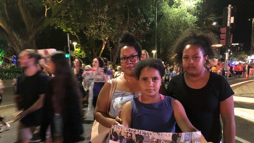 Three women hold a poster while hundreds march by
