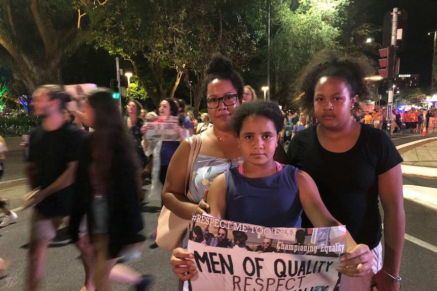 Three women hold a poster while hundreds march by