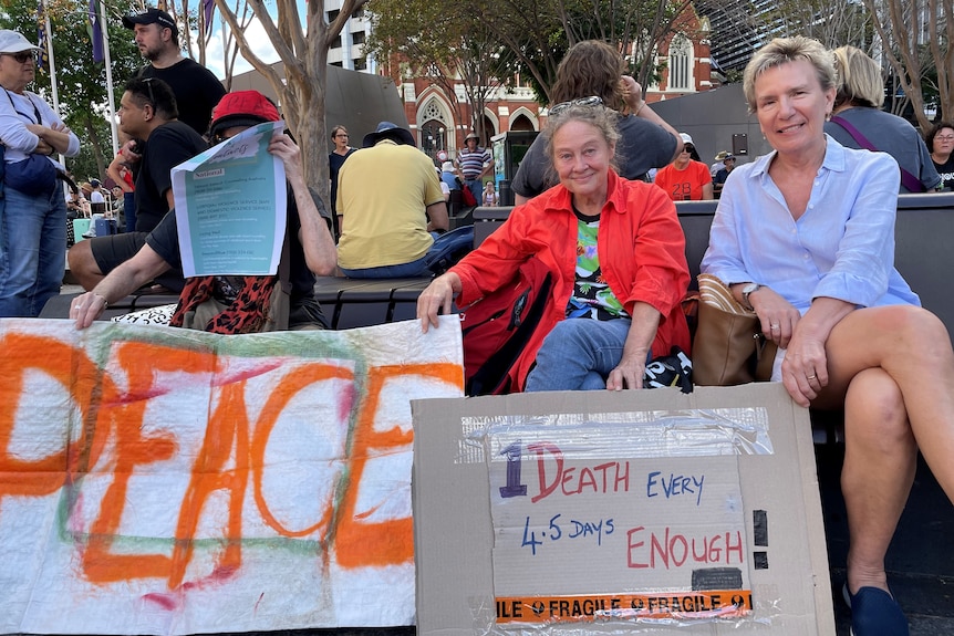 Three people sit on a bench with two signs in front of them. One say peace, the other says one death evey 4.5 days is enough.
