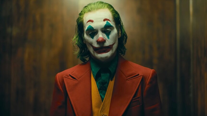 The Joker, with clown face paint, standing in front of a wood-panelled wall.