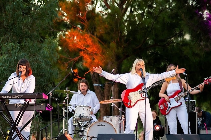 Four musicians in white boiler suits play on an outdoor stage, the frontwoman with her arms spread wide as she sings.