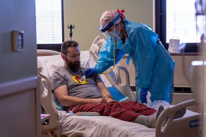 A woman in scrubs and full PPE leaning over a man sitting in a hospital bed with oxygen cannula under his nose