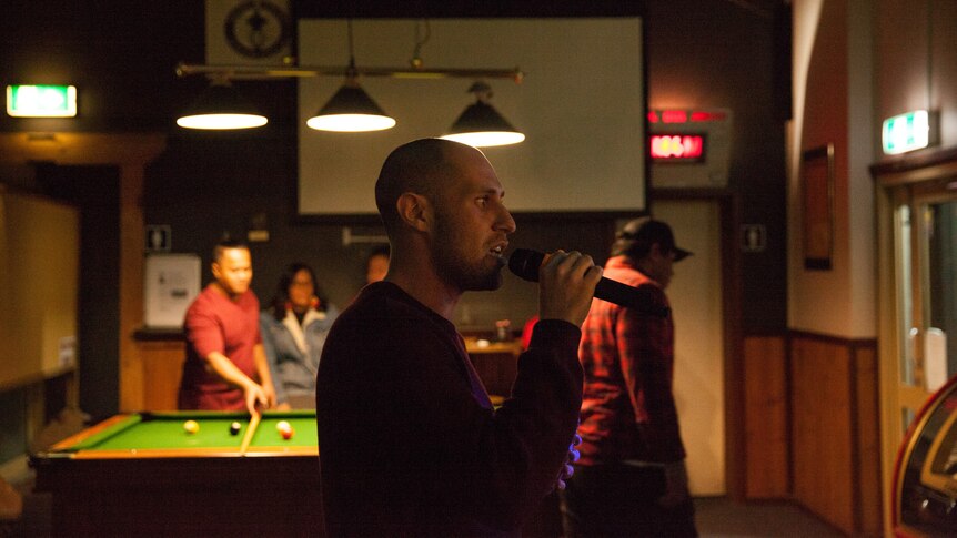 A young man holds a microphone to his mouth, as others play pool nearby.