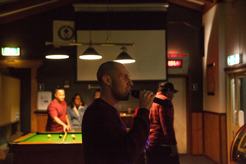 A young man holds a microphone to his mouth, as others play pool nearby.