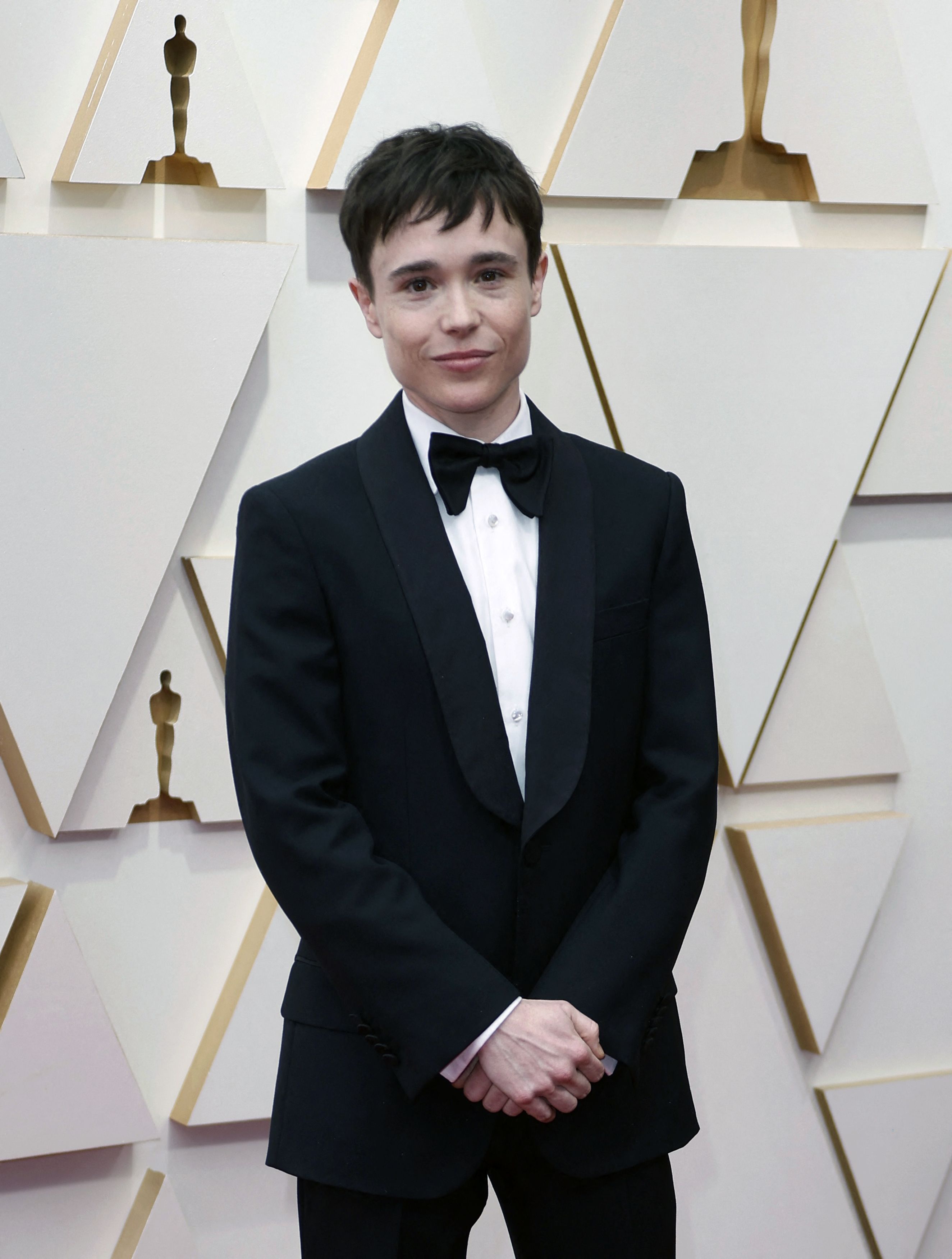 Elliot Page stands in front of a white media wall wearing a black suit with a white shirt and black bow tie.