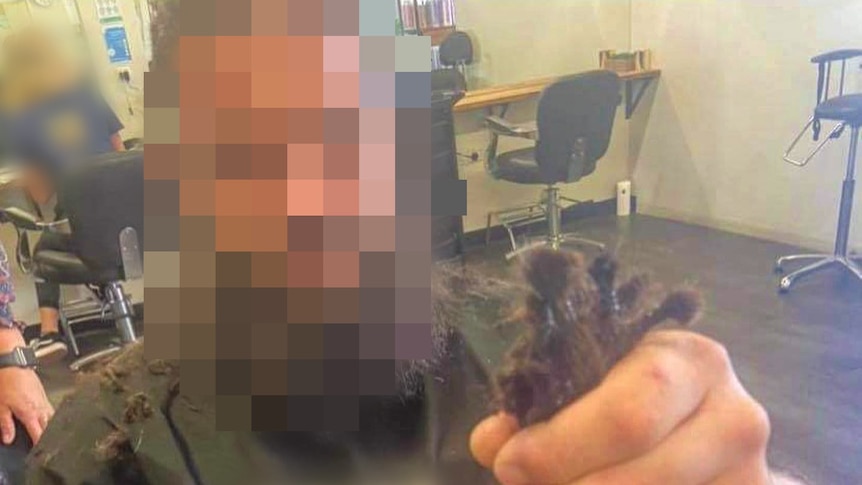 A man in a hair salon chair holds up dreadlocks that have been cut off.