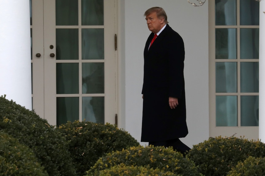 Donald Trump in a coat and tie walks along the outside of the White House