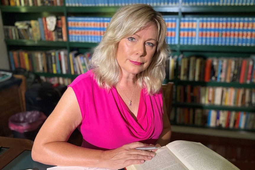 A blonde woman in a bright top sits, with a book open in front of her, in what appears to be a private library.