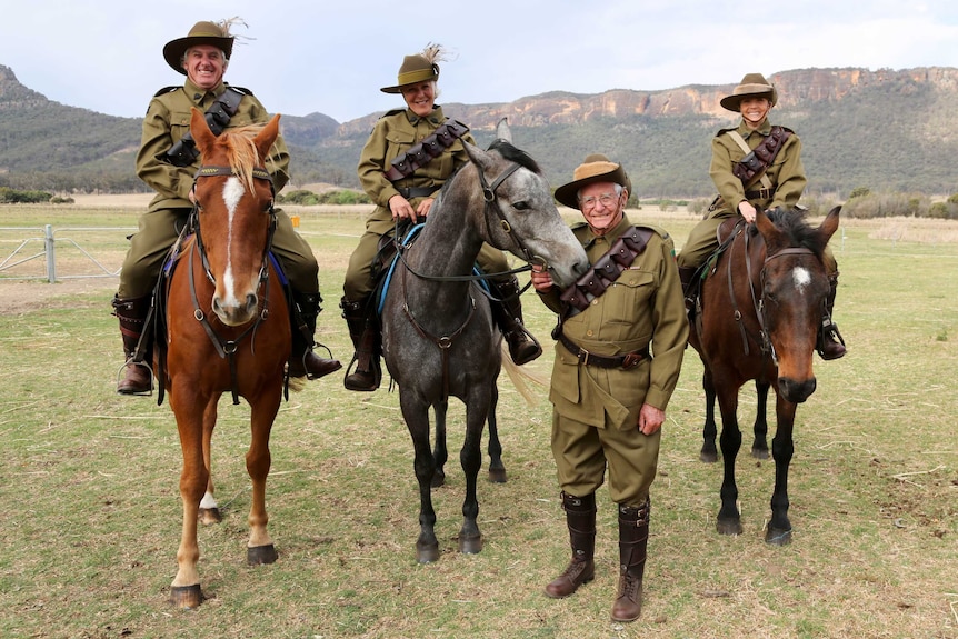 A man stands with three people on horseback. All are dressed in military uniforms.