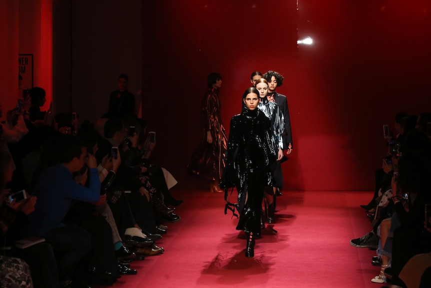 A group of fashion models walk in a line on a red carpeted catwalk.