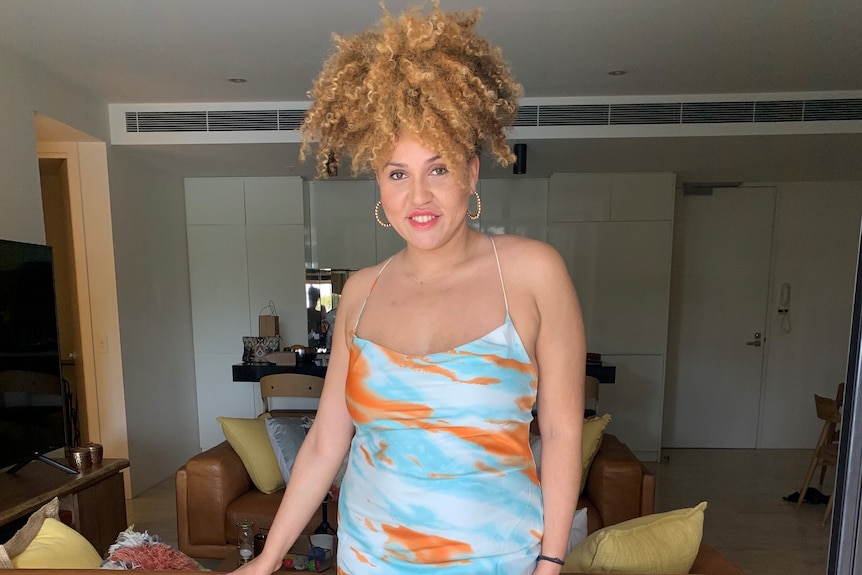A person looks to the camera while smiling. They wear a blue and orange dress and their Afro hair is tied up in a puff.