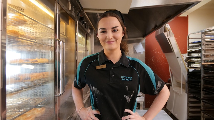 Girl working in a bakery kitchen smiling with flour on hands and on shirt with hands on hips. Pies in warming ovens behind.