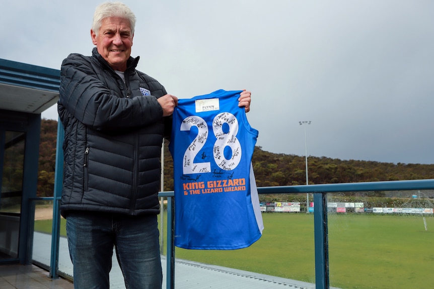 Man with white hair wearing puffer jacket holding blue football jumper on balcony overlooking football ground