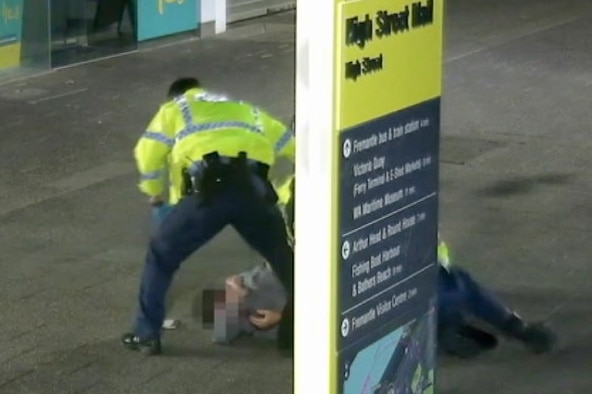 A man lies hunched over on the ground in a mall with a hand over his face and a police officer standing over him.