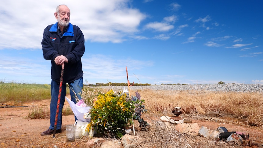 An older man with a beard leans on a walking stick and looks at crosses and flowers near a rail line.