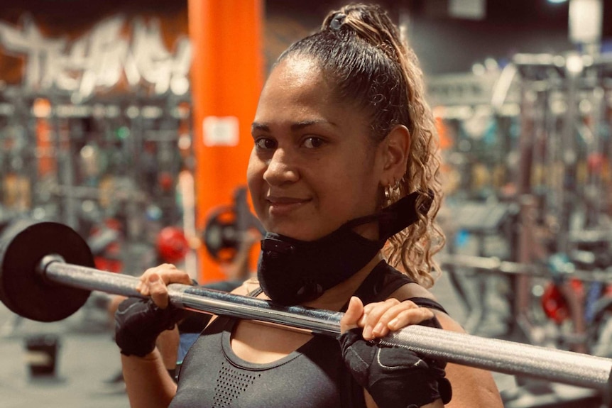 A woman smiles as she looks to the camera while holding a bar bell. She has gloves on and a black mask around her neck.