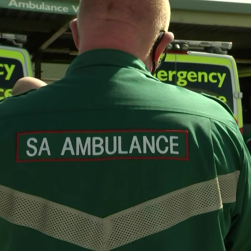A paramedic stands with his back to the camera in front of ambulances and a man wearing a suit