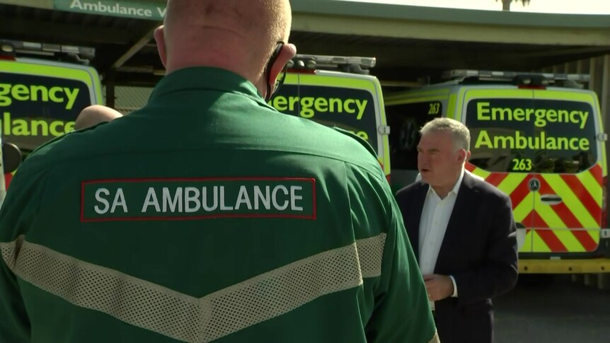 A paramedic stands with his back to the camera in front of ambulances and a man wearing a suit