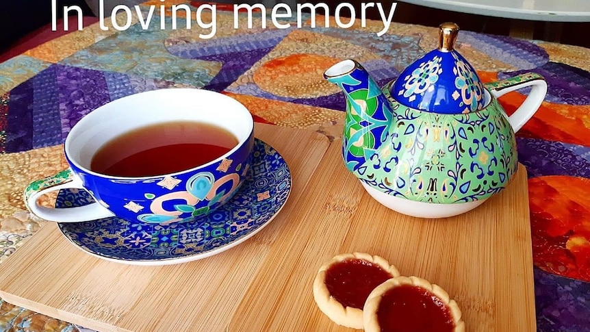 A decorative tea cup and tea pot on a wooden board with two jam tarts next to them.