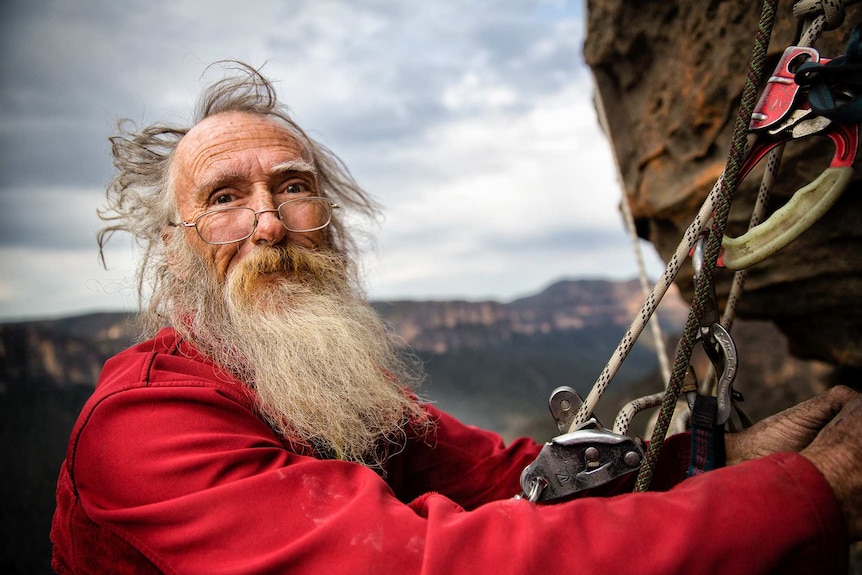 a bearded man hanging off a mountain side smiling