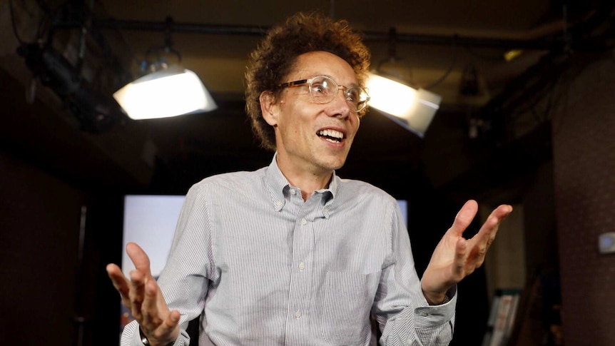 Malcolm Gladwell throws his hands in the air and smiles in a photo in a studio