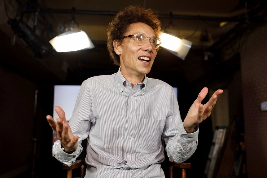 Malcolm Gladwell throws his hands in the air and smiles in a photo in a studio