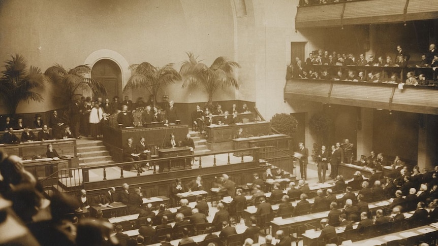 A sepia tone image of a large hall filled with suited men.