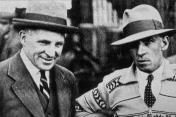 A black and white image of Johnnie Hoskins wearing a sweater and hat next to another man. 